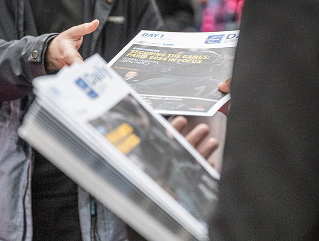 A person handing out the daily news, the Milipol Paris newspaper, to another person at the entrance to the exhibition
