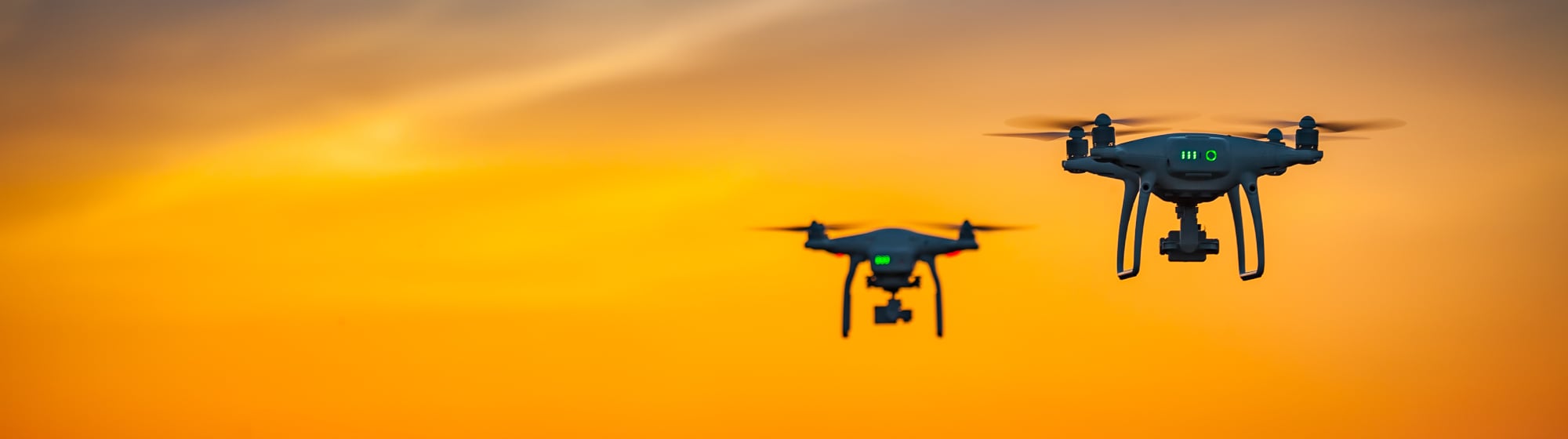 Two drones flying at sunset, silhouetted against the orange sky, symbolizing aerial surveillance.