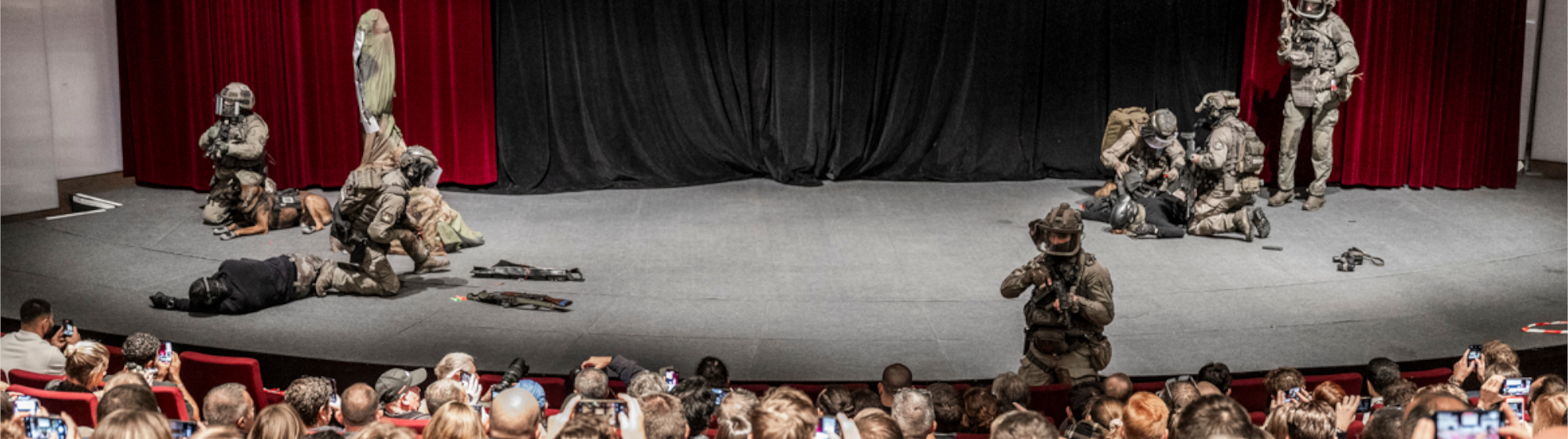 Live tactical demonstration by soldiers in combat gear on an auditorium stage, observed by an audience taking pictures.