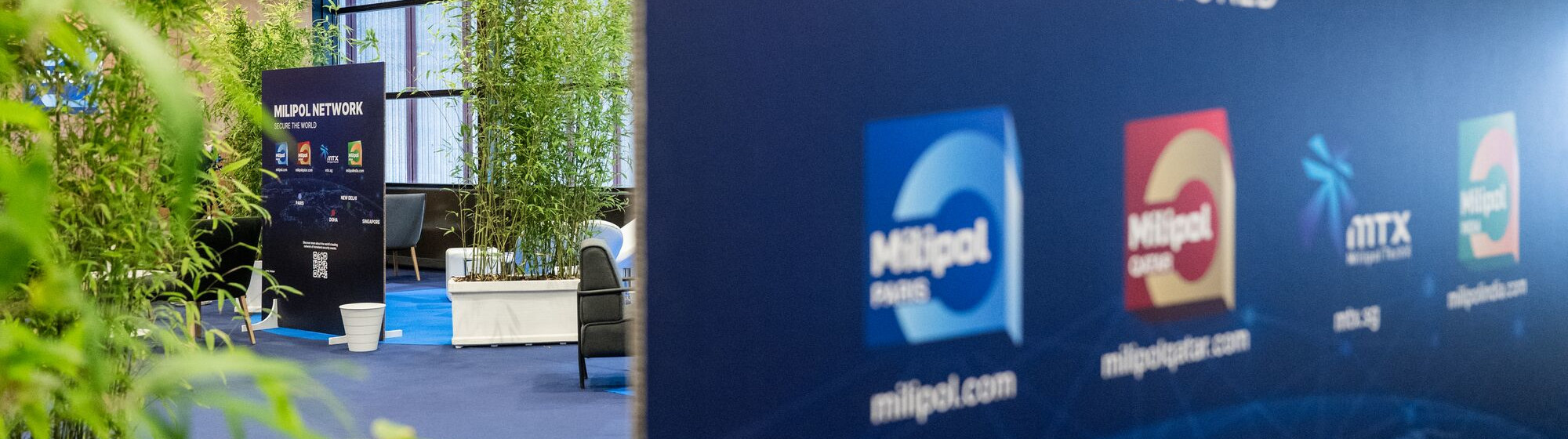 Espace lounge avec des plantes vertes et une grande bannière Milipol Network affichant les logos des événements Milipol à Paris, au Qatar, en Inde et à Singapour