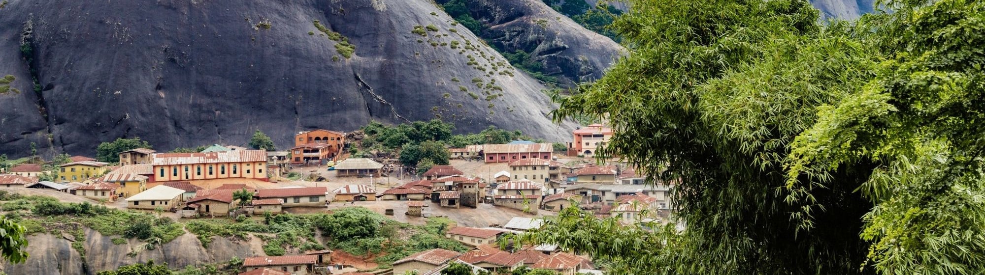 Panoramic view of a Nigerian village surrounded by rocky mountains with greenery, under a cloudy sky.
