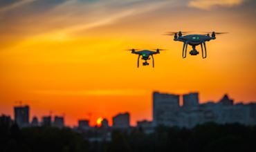 Two drones flying above a city at sunset, silhouetted against the orange sky, symbolizing aerial surveillance.