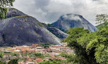 Vue panoramique d’un village nigérian entouré de montagnes rocheuses couvertes de végétation, sous un ciel nuageux.