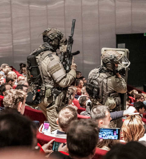 Simulated GIGN intervention in a crowded auditorium during Milipol Paris, with two armed tactical officers standing among the audience