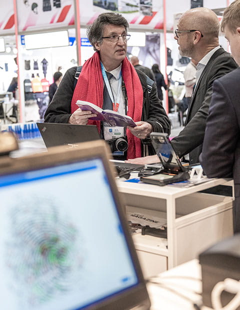 Un visiteur et un exposant échangent ensemble sur un stand. Un écran d'ordinateur projete une emprunte digitale sur son écran