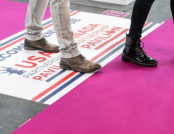Zoom sur les jambes de deux visiteurs machant dans les allées, avec signalétique au sol indiquant le pavillon USA