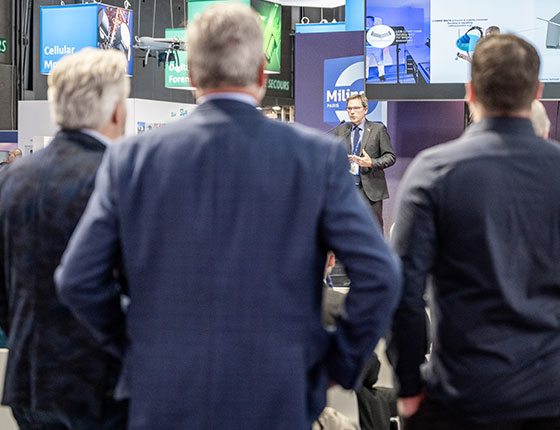 3 hommes debout assistent à l'intervention d'un exposant sur l'innovation stage
