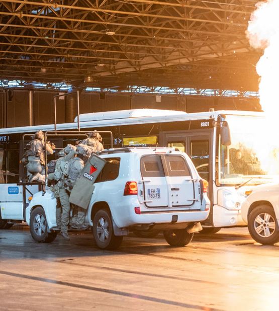 GIGN intervention simulation at Milipol Paris, with tactical officers boarding a bus using an assault vehicle, under a controlled explosion