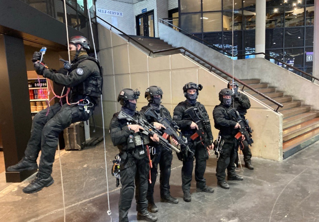Un groupe de policiers de la brigade d'intervention (BI/DOPC) armés, en uniformes noir se tiennent dans le hall avec un dernier membre descendant en rappel à l'aide d'une corde depuis le toit