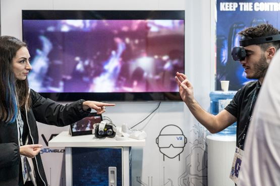 Une femme guide un homme équipé d’un casque de réalité virtuelle, devant un écran diffusant des images floues dans un espace d’exposition technologique.
